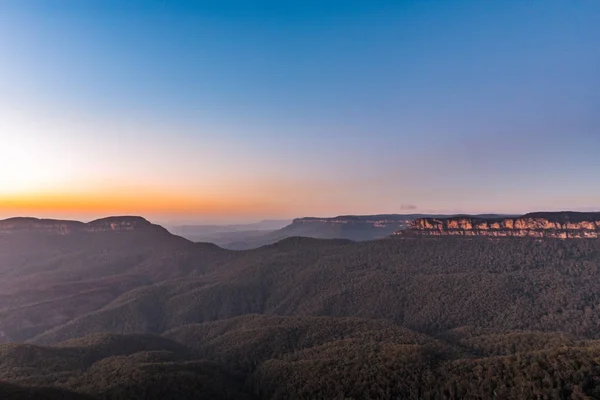 The sun rises slowly over the dense forests of the Blue Mountains in NSW, Australia. The sun creates a beautiful orange to blue gradient.