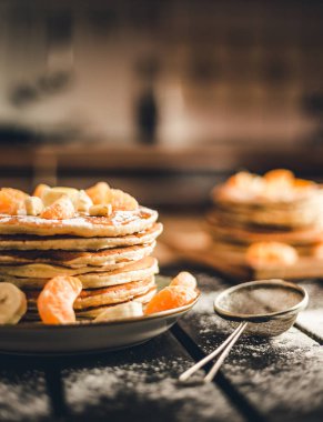 Dikey fotoğraf - muz ve portakal ahşap tahta üzerinde altın krep yığını pudra şekeri ile kaplı. Ahşap masa üzerinde elek ile akçaağaç şurubu ile Amerikan krep yığını.