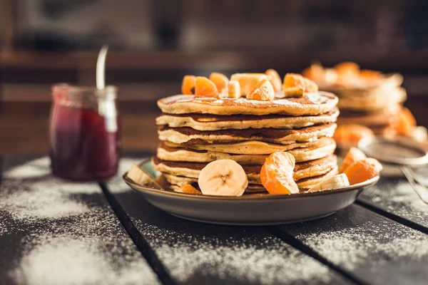 Stack of golden pancakes with bananas and oranges on wooden board covered with caster sugar. Heap of american pancakes with maple syrup and a glass of jam in kitchen.