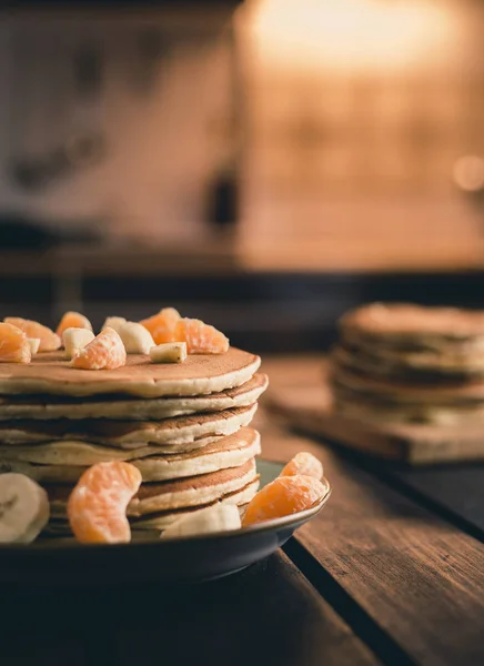 Vertical photo - stack of golden pancakes with bananas and oranges on wooden board covered with caster sugar. Heap of american pancakes with maple syrup with sieve on wooden table -light on background