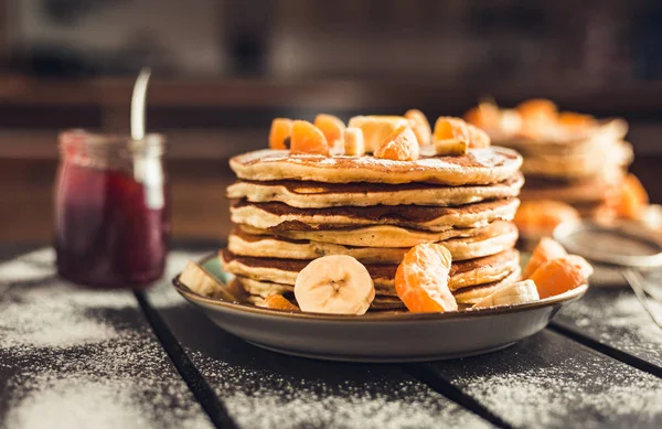 Stack of golden pancakes with bananas and oranges on wooden board covered with caster sugar. Heap of american pancakes with maple syrup and a glass of jam in kitchen.