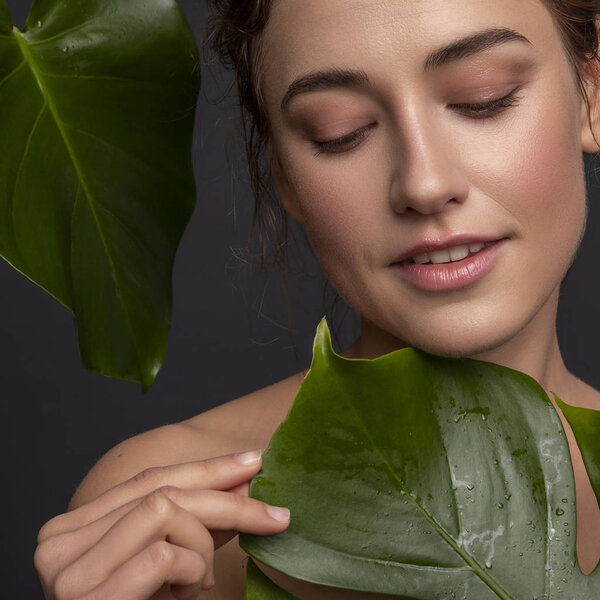Cute ordinary young woman posing with monstera tropical leafs. Fresh clean skin with flawless texture. Skin care beauty cosmetology concept. Space for text.
