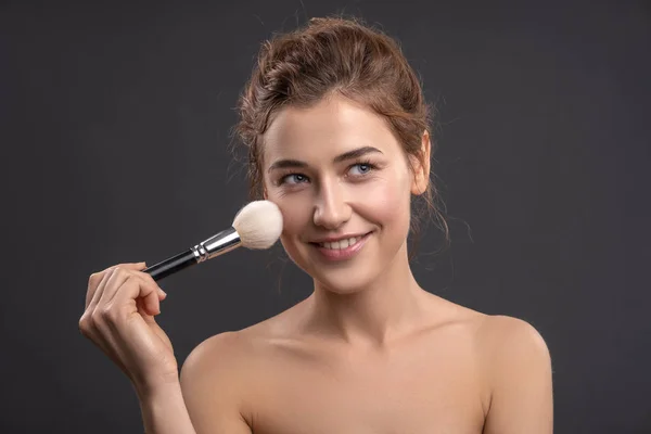Beautiful brunette caucasian young woman prepare herself, applying powder on her cheeks with a brush. Clean, fresh, natural, flawless skin. Soft smile on her face. Close up on a neutral background