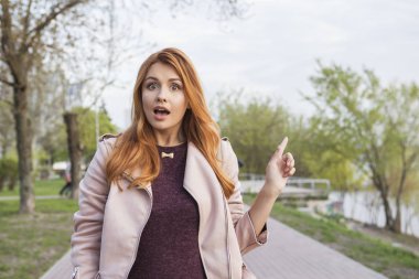 Kızıl saçlı seksi artı bir parkta yürüyen boyutu kadın. Soğuk havada deri ceket giyiyor. Duygusal portre. Kopya alanı üzerinde parmaklarını işaret ediyor