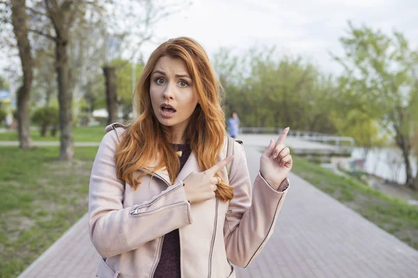 赤い髪のセクシープラスサイズの女性が公園を歩いている 寒い天候の彼女は革のジャケットを着ている 感情的な肖像画彼女はコピースペースに指を向けます — ストック写真