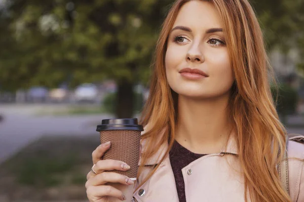 赤い髪のセクシープラスサイズの女性が公園を歩いている 寒い天候の彼女は革のジャケットを着ている 彼女は カップからコーヒーを飲みます 背景に見事な夕日 スペースのコピー — ストック写真