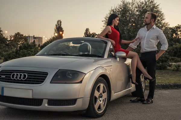 Gorgeous sexy copule of man and woman on a vacation car trip in summer day. Luxury grey sport car. Sexy fit brunette woman and man in evening outfit. Night portrait.