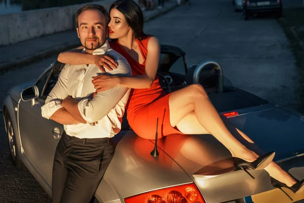 Gorgeous sexy copule of man and woman on a vacation car trip in summer day. Luxury grey sport car. Sexy fit brunette woman and man in evening outfit. Night portrait.