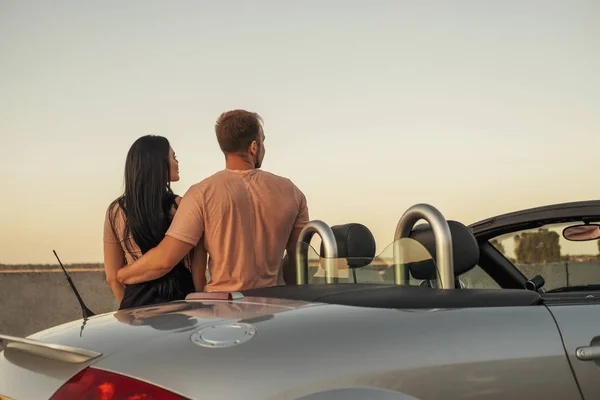 Gorgeous sexy copule of man and woman on a vacation car trip in summer day. Luxury grey sport car. Sexy fit brunette woman and man in casual outfit. Sunset.