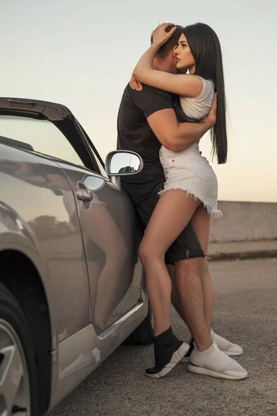 Gorgeous sexy copule of man and woman on a vacation car trip in summer day. Luxury grey sport car. Sexy fit brunette woman and man in casual outfit. Sunset.