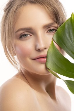 Sevimli gülümseyen sarışın beyaz kadın taze, temiz, sağlıklı, kusursuz bir cilt. Çıplak, gündüz makyaj rahat saç modeli ile. Portre tropikal monstera yaprak modeli yüz yakınındaki bir beyaz zemin üzerine.