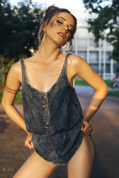 Cute sexy woman in jeans overall outfit with glitter freckles make up and hair buns. Summer evening in the city. Sensual posing.