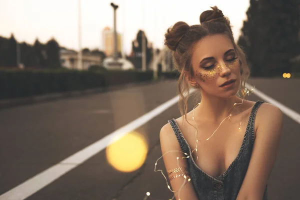 Cute sexy woman in jeans overall outfit with glitter freckles make up and hair buns. Summer evening in the city. Sensual posing in garland golden lights.