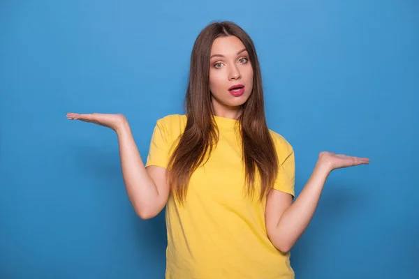 青い背景に黄色のTシャツでポーズ長い髪を持つかわいいブルネットの女性 感情的な肖像画彼女は何を選べばいいのだろうかと疑いながら手を握っている — ストック写真