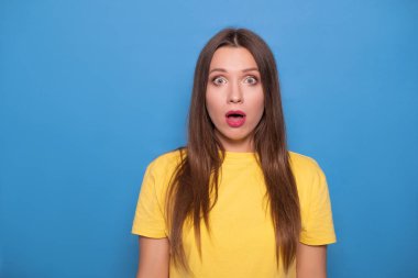 Şirin esmer kadın uzun saçlı mavi bir arka plan üzerinde sarı tişört poz. Duygusal portre. O şok, hayret