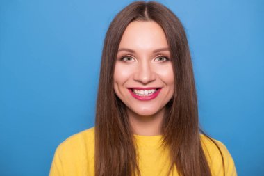 Mavi bir arka plan üzerinde sarı t-shirt poz uzun saçlı sevimli esmer kadın. Duygusal portre. Kusursuz beyaz dişleri ile mutlu bir şekilde gülümsüyor.