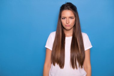 Şirin esmer kadın uzun saçlı mavi bir arka plan üzerinde beyaz t-shirt poz. Duygusal portre. O mutsuz yüz yapmak
