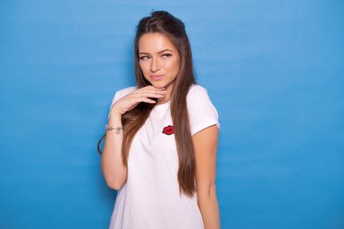 Şirin esmer kadın uzun saçlı mavi bir arka plan üzerinde beyaz t-shirt poz. Duygusal portre. O flört ediyor