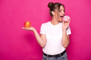 Seksi model pembe bir arka plan üzerinde beyaz tişört içinde şık saç modeli ile. Duygusal portre. O şok, hayret, iyi ve kötü gıda arasında seçmek için bağlama: elma veya çörek?