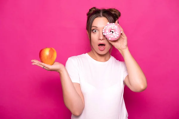Sexy model with stylish hairstyle in white t-shirt on a pink background. Emotional portrait. She shocked, amazed, tying to choose between good and bad food: apple or donut?