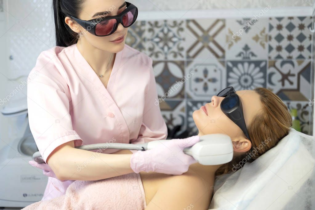 Woman doctor cosmetologist removing hair from a woman client body using laser epilator. They in a spa center.