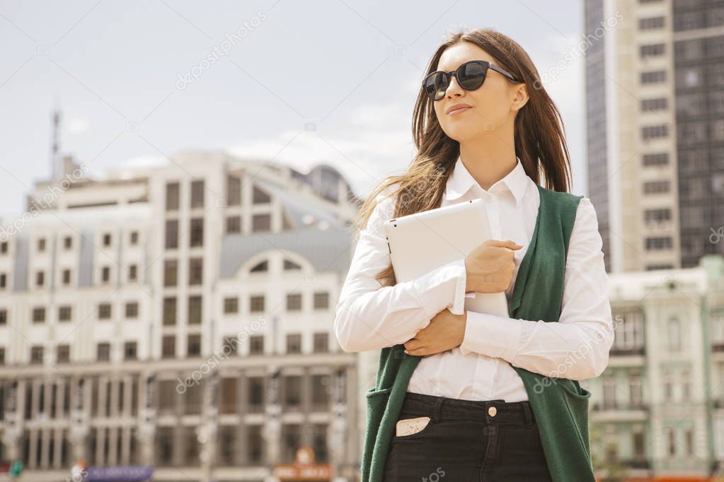 Beautiful brunette business woman in white blouse and green jacket working on a tablet in her hands outdoors. Freelancer in european city. Space for text.