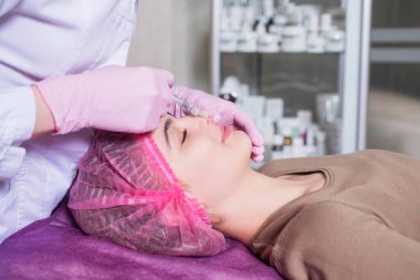 Kozmetik doktoru kliniğinde bir kadına iğne yapıyor. Parıltı ve maske takıyor ve şırıngayı iğneyle tutuyor..