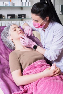 Kozmetik doktoru kliniğinde bir kadına iğne yapıyor. Parıltı ve maske takıyor ve şırıngayı iğneyle tutuyor..