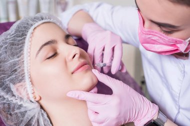 Kozmetik doktoru kliniğinde bir kadına iğne yapıyor. Parıltı ve maske takıyor ve şırıngayı iğneyle tutuyor..