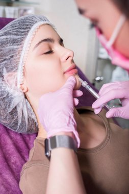 Kozmetik doktoru kliniğinde bir kadına iğne yapıyor. Parıltı ve maske takıyor ve şırıngayı iğneyle tutuyor..