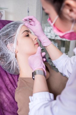 Kozmetik doktoru kliniğinde bir kadına iğne yapıyor. Parıltı ve maske takıyor ve şırıngayı iğneyle tutuyor..