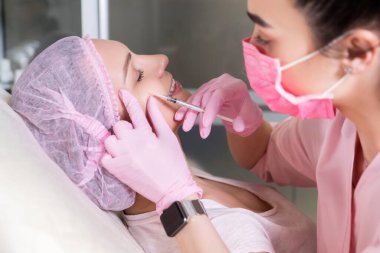Kozmetik doktoru kliniğinde bir kadına iğne yapıyor. Pembe parıltı ve maske takıyor ve şırıngayı iğneyle tutuyor..