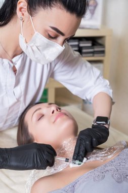 Kozmetik doktoru kliniğinde bir kadına iğne yapıyor. Siyah parıltı ve maske takıyor ve şırıngayı iğneyle tutuyor..