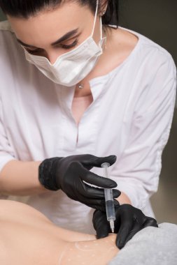 Kozmetik doktoru kliniğinde bir kadına lipolitik enjeksiyon yapıyor. Siyah parıltı ve maske takıyor ve şırıngayı iğneyle tutuyor..