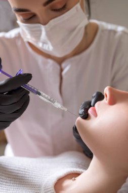 Kozmetik doktoru kliniğinde bir kadına iğne yapıyor. Siyah parıltı ve maske takıyor ve şırıngayı iğneyle tutuyor..