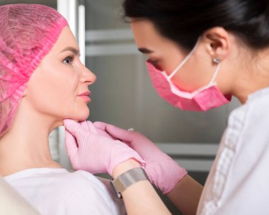 Kozmetik doktoru klinikteki kadın bir müşteriye iğne yapıyor. Maske ve eldiven takıyor..