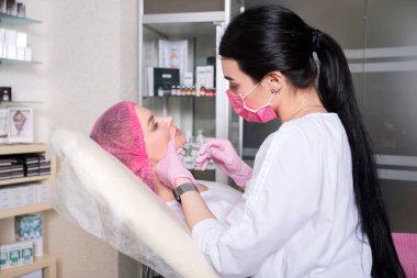 Kozmetik doktoru klinikteki kadın bir müşteriye iğne yapıyor. Maske ve eldiven takıyor..
