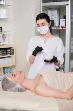 Kozmetik doktoru kliniğinde bir kadına enzim tedavisi uyguluyor. Parıltı ve maske takıyor..