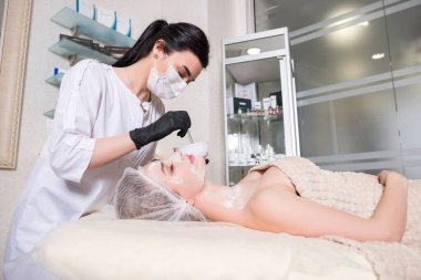 Kozmetik doktoru kliniğinde bir kadına enzim tedavisi uyguluyor. Parıltı ve maske takıyor..