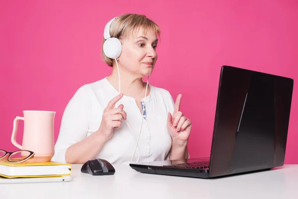 Old woman in her 60s works on computer, wearing headphohes. Laptop on white table and Pink background. She participate in online conference or webinar, teach others.