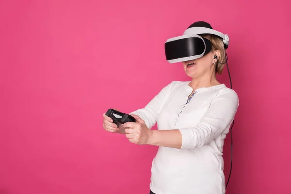 Elderly in 60s woman plays in VR helmet. She stands in white blouse on pink background and looks happy.