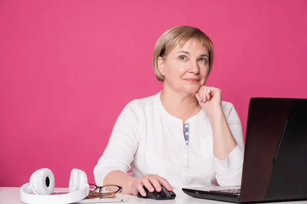 Old woman in her 60s works on computer, wearing headphohes. Laptop on white table and Pink background. She thoughtful, thinking about something.