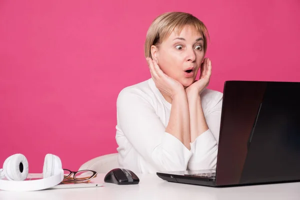 Old woman in her 60s works on computer, wearing headphohes. Laptop on white table and Pink background. She amazed, surprised abouth the news she recieved.