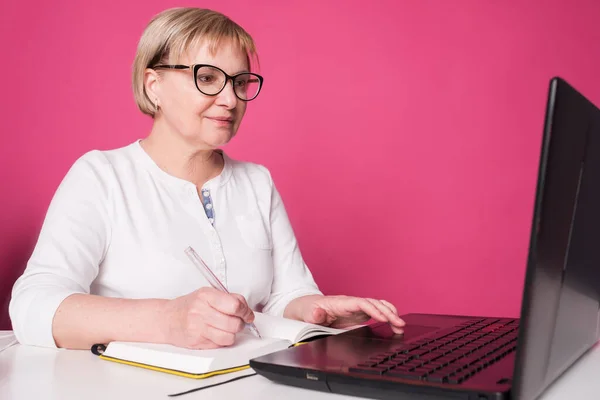 Old woman in her 60s works on computer, wearing headphohes. Laptop on white table and Pink background. She make notes on her notebook with a pen