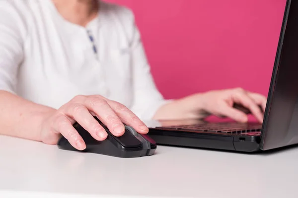 Close up f an elderly woman hands on mouse and laptop keyboard. She plays computer games or searching in web, chatting in social media sites. White table and pink background