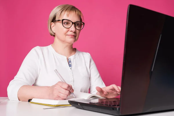 Old woman in her 60s works on computer, wearing headphohes. Laptop on white table and Pink background. She make notes on her notebook with a pen