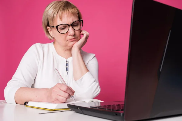 Old woman in her 60s works on computer, wearing headphohes. Laptop on white table and Pink background. She sits thoughtful, unhappy, a bit sad