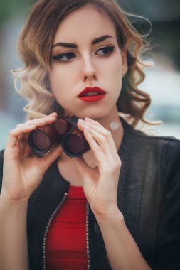 Güzel esmer seksi ajan (katil veya polis) Deri ceketli ve elinde dürbün olan kırmızı lüks elbiseli kadın Avrupa 'daki birini izliyor..