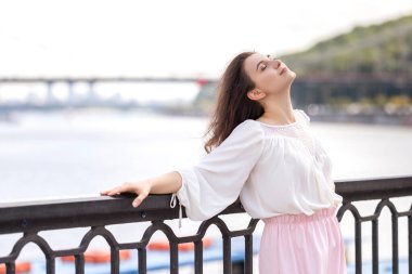 Romantik kıyafetli genç bir kadın nehir kenarındaki Avrupa sokaklarında. Yaz güneşi altında tek başına dinleniyor. Metin için boşluk.