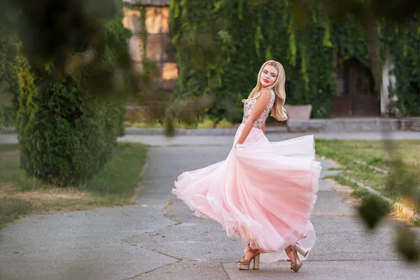 Blonde woman with make up in evening dress at park. The skirt of her dress fly on a wind. Sensual fashion portrait with a sunset light.